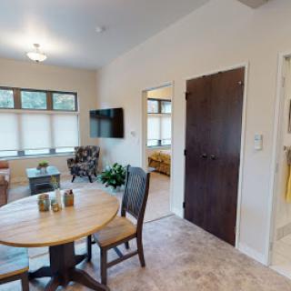 A cozy living space featuring a dining area with a round table and chairs, a television mounted on the wall, and natural light streaming through the windows.