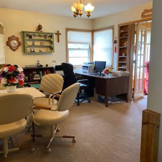 A cozy home office featuring a desk with a computer, comfortable chairs around a table, shelves with decor, and large windows providing natural light.