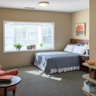 A cozy bedroom with a large window allowing natural light, featuring a neatly made bed and comfortable seating.