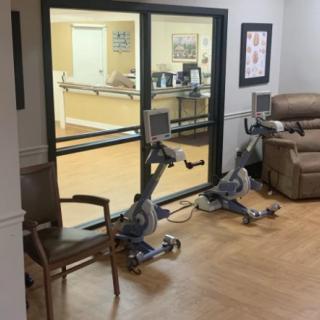 A healthcare facility room featuring two stationary bikes and chairs near a glass window.
