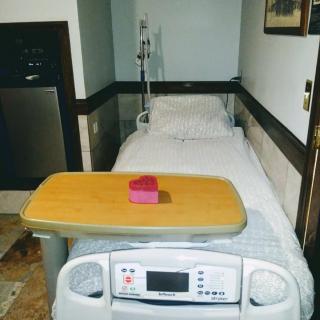 A hospital bed with an IV stand in a patient room, featuring a tray table with a small pink item.