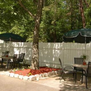 A peaceful outdoor patio area with tables and chairs, shaded by umbrellas and surrounded by trees.