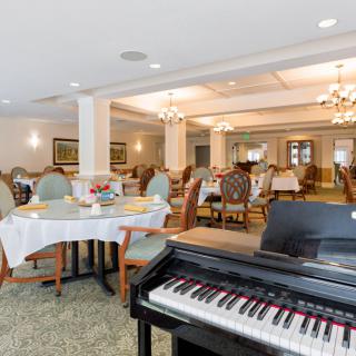 A spacious dining room with elegant decor features a piano in the foreground and several tables and chairs set for dining.