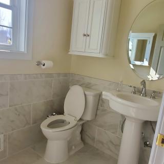 A clean and bright bathroom featuring a toilet, pedestal sink, round mirror, and a wall-mounted cabinet.