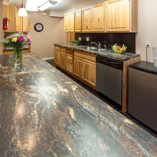 A modern kitchen with wooden cabinets and a marble countertop, featuring a sink, flowers, and a fruit bowl.