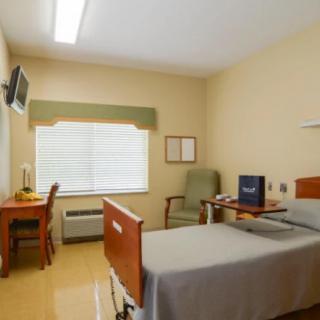 A clean and organized hospital room with a bed, desk, chair, and television.