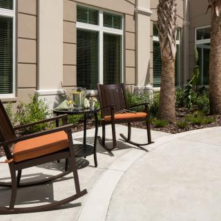 A cozy patio area with two rocking chairs and a table, surrounded by greenery and palm trees.