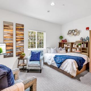 A cozy bedroom with modern decor, featuring a comfortable bed, a chair, and shelves filled with books and decorative items.