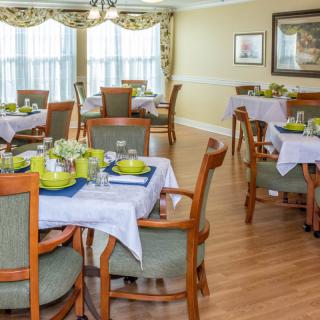 A well-decorated dining room with multiple tables and chairs, featuring neatly arranged table settings and a bright wood floor.