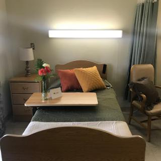 A cozy hospital room featuring a single bed with decorative pillows, a nightstand, and a chair.