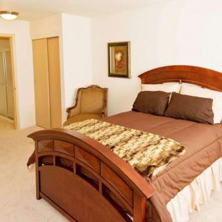 A well-lit bedroom featuring a wooden bed with brown bedding and a decorative throw, complemented by a classic chair and wall art.