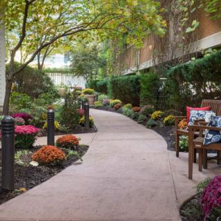 A serene garden pathway lined with colorful flowers and benches under a canopy of trees.
