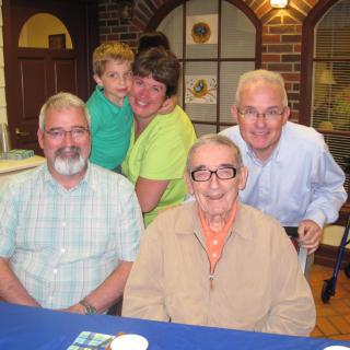 A family group photo with three adults, an elderly person, and a child smiling indoors.