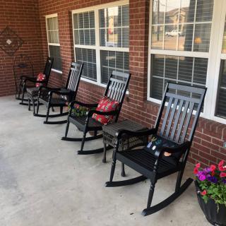 A row of black rocking chairs with cushions on a porch with a brick wall and windows, accompanied by a flower pot.