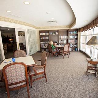 A cozy library room with bookshelves, tables, and chairs by a large window.