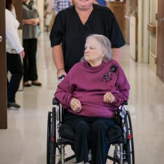 A caregiver assists an elderly woman in a wheelchair through a hospital corridor.