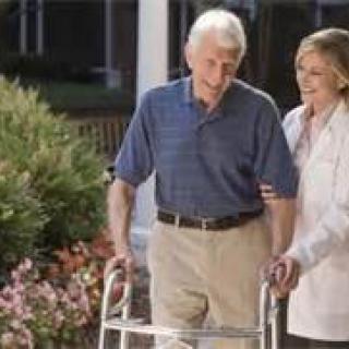 An elderly man using a walker is assisted by a caregiver outdoors.