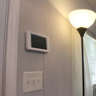 A smart thermostat mounted on a wall beside light switches and a floor lamp in a well-lit room.