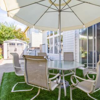 A sunny backyard patio with a glass table, chairs, and an umbrella next to a garden shed.