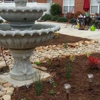 A landscaped garden featuring a tiered fountain surrounded by mulch and young plants, with patio furniture in the background.