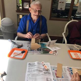 An elderly man engaged in a craft activity, smiling as he works at a table in a well-lit room.
