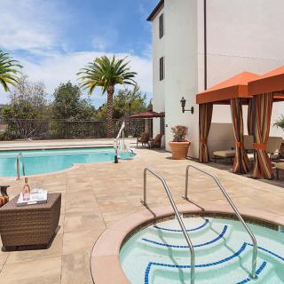 An inviting outdoor pool area with a hot tub, cabanas, and palm trees under a clear blue sky.
