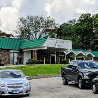 A view of a building with a green roof, surrounded by parked cars and lush trees under a cloudy sky.