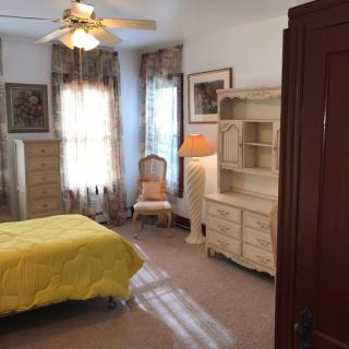 A cozy bedroom with vintage-style furniture, featuring a bed with a yellow cover, a dresser, and natural light streaming through the windows.