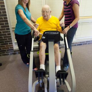 An elderly man receiving assistance with physical therapy exercises on a specialized machine.