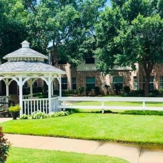 A beautiful garden with a white gazebo, surrounded by lush green grass and trees, adjacent to a building.