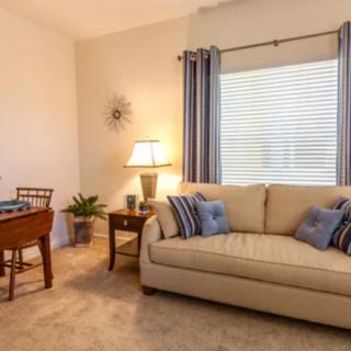 A cozy living room featuring a sofa with throw pillows, a table with a lamp, and large window with curtains.