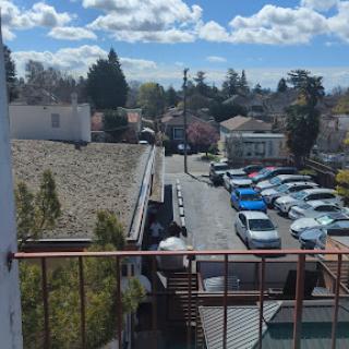 A view from a balcony overlooking a quiet street with parked cars and fluffy clouds in the sky.