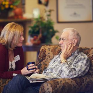 A caregiver engages in a warm conversation with an elderly man in a comfortable indoor setting.