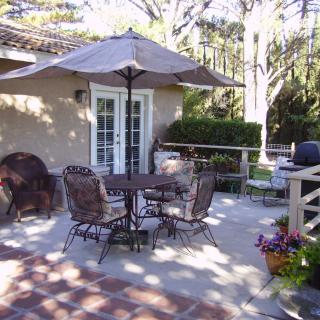 A sunny patio with outdoor furniture, including a table and chairs under an umbrella, surrounded by greenery.