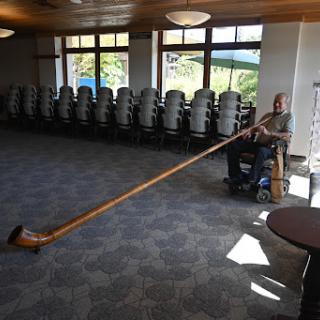 A musician plays a long wooden alphorn inside a spacious hall.