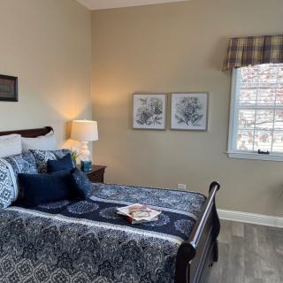 A cozy bedroom featuring a patterned bedspread, two framed artworks above the bed, a bedside lamp, and a window with plaid curtains.