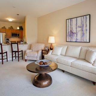 A cozy living room with a cream sofa, armchair, and modern coffee table, adjacent to an open kitchen.