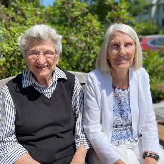 Two elderly women sitting on a bench outdoors, enjoying the sunshine and smiling.