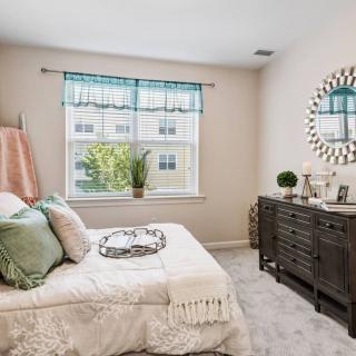 A cozy bedroom with a neatly made bed, decorative pillows, and a dresser topped with plants and decor, illuminated by natural light from the window.
