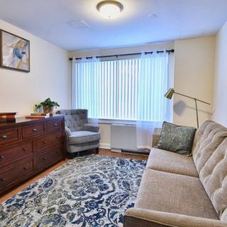 A cozy living room with a sofa, armchair, and patterned rug, featuring a window with blinds and a standing lamp.
