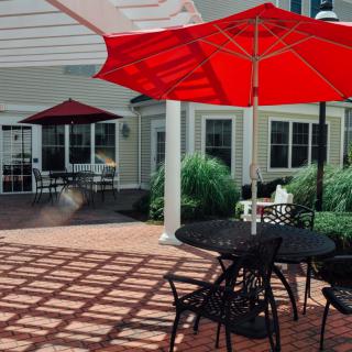 A sunny patio with red umbrellas, providing shade over tables and chairs on a brick floor.