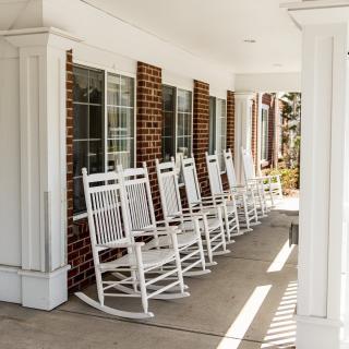 A row of white rocking chairs lined up on a porch with a brick wall and windows.