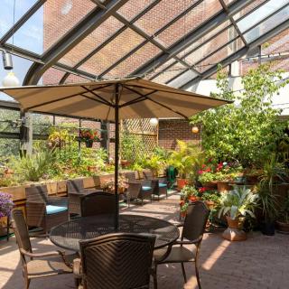 A cozy indoor greenhouse with a glass roof, filled with lush plants and patio seating under an umbrella.