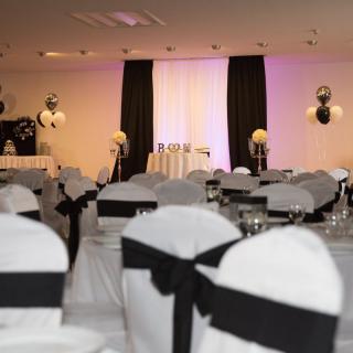 An elegant banquet hall set up with a black and white theme, featuring decorated chairs and ambient lighting.