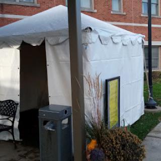 A white tent set up in a public area, featuring a chair and a nearby trash bin.