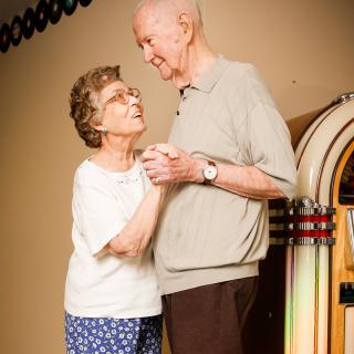 An elderly couple shares a joyful dance next to a jukebox.