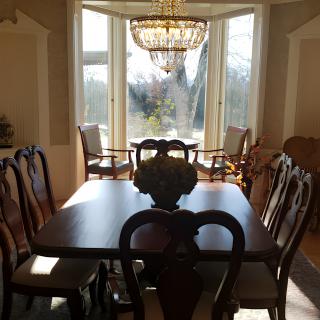 A dining room with a wooden table and chairs, illuminated by a chandelier, showcasing a bay window with natural light.