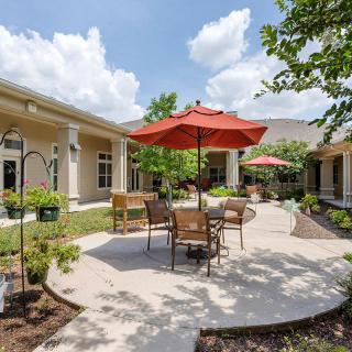 A sunny outdoor patio area with tables, chairs, and red umbrellas surrounded by landscaped gardens.