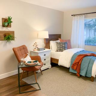 A cozy and modern bedroom featuring a comfortable bed with colorful pillows and throws, a stylish chair with a small table, and soft lighting from a bedside lamp.