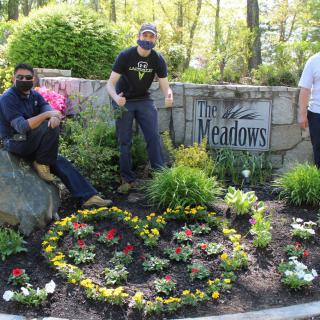 Three individuals posing in a garden setting with colorful flowers and a sign reading 'The Meadows' in the background.